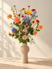 Apricot Garden zijden boeket met abrikooskleurige, oranje, lila en blauwe bloemen in een matte zandkleurige vaas.